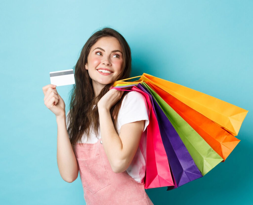 young satisfied woman smiling, showing plastic credit card and holding shopping bags, buying with contactless payment, standing over blue background